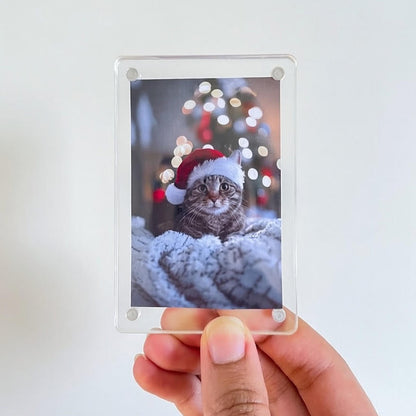 Person holding a photo of a cat wearing a Santa hat with a blurred Christmas tree in the background.